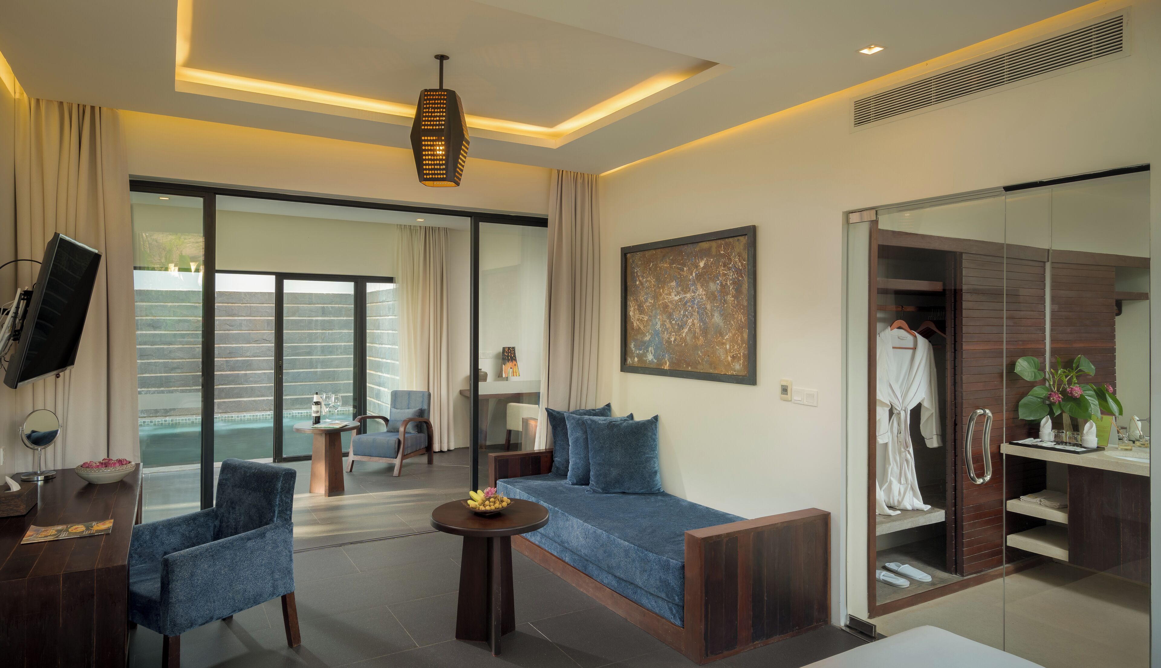 A modern hotel room with a sofa, chairs, a small table, a TV, a painting, and a bathroom visible through a glass door.