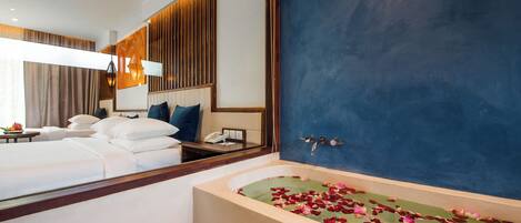 A hotel room with a large bed, a bathtub with rose petals, a mirror, and a nightstand with a phone.