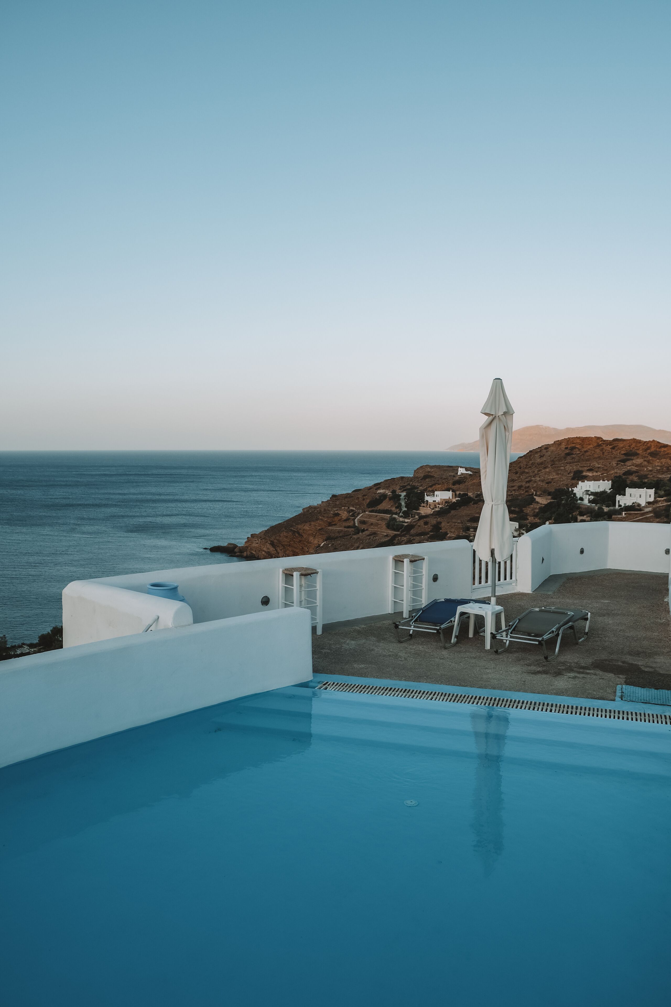 Un espace piscine doté de chaises longues et d’un parasol, offrant une vue sur un paysage côtier avec des bâtiments blancs et des collines.
