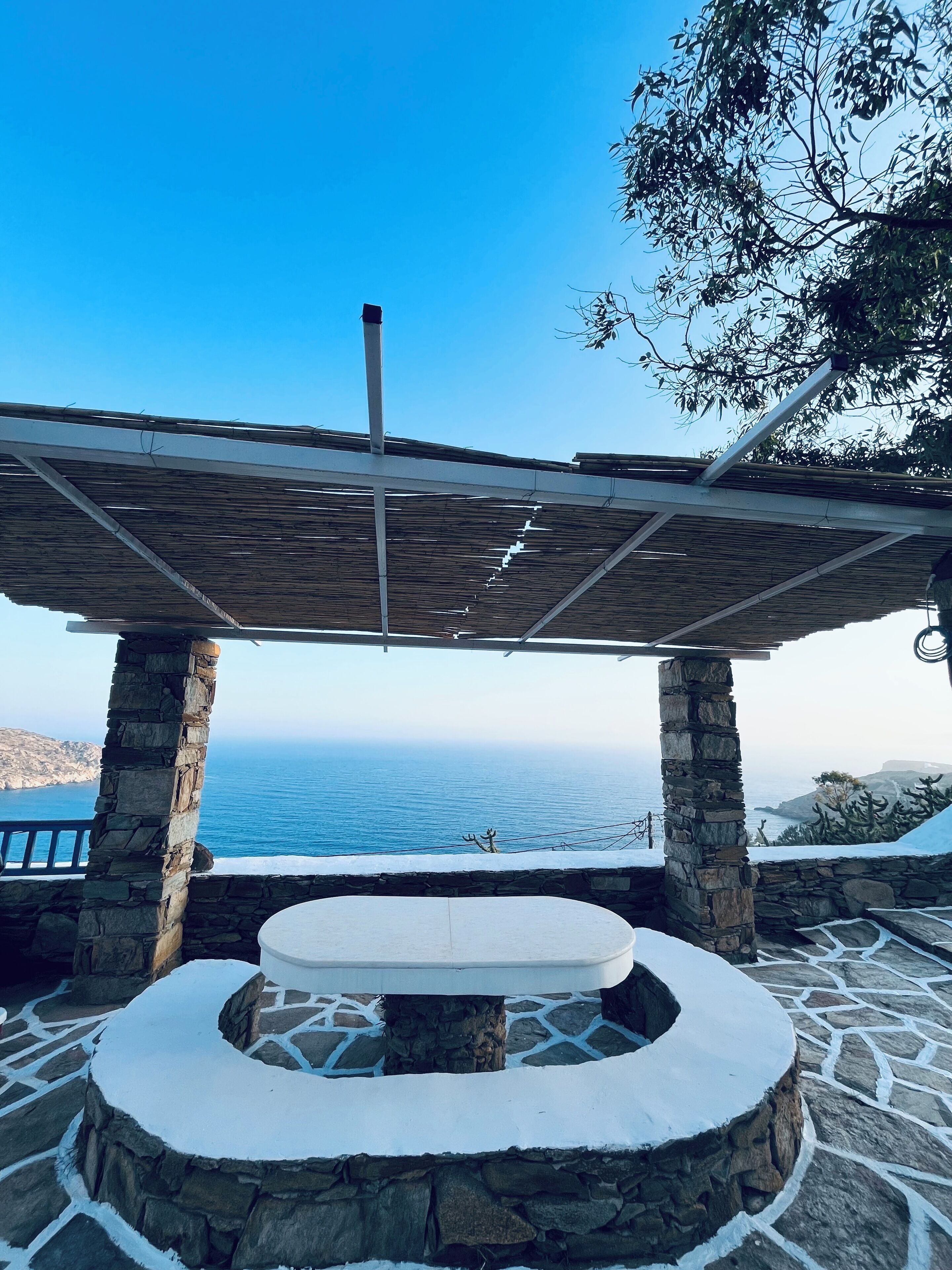 A stone patio with a circular seating area, a pergola overhead, and a view of the sea.