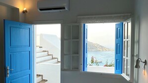 A bedroom with a bed, a blue door, a window with an open view, and an air conditioning unit. - Petradi Hotel (Ios)