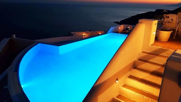 A curved infinity pool with blue lighting, overlooking a coastal landscape at sunset. - Petradi Hotel (Ios)