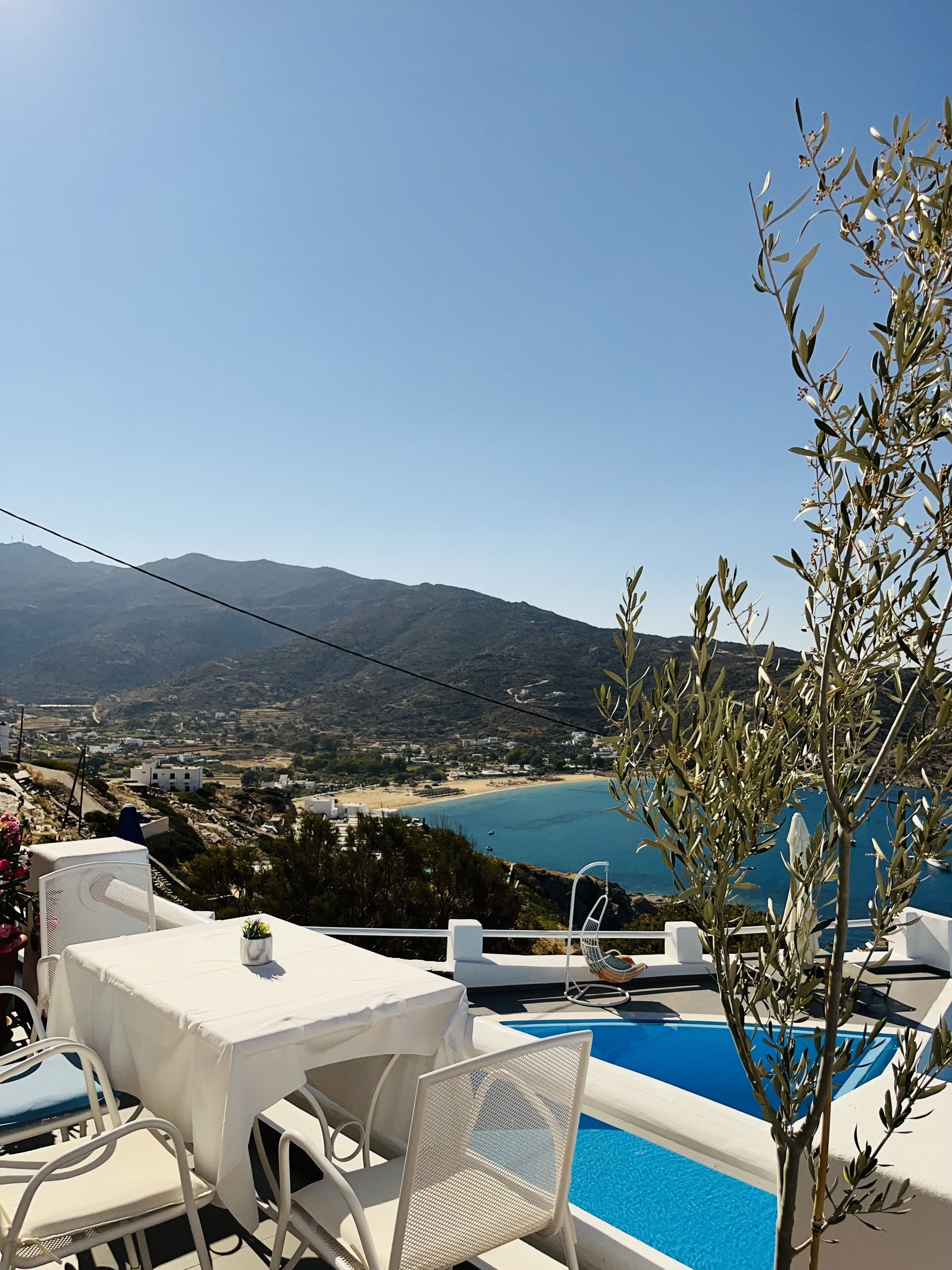 Ein Poolbereich mit weißer Möbel, Blick auf einen Strand und Berge im Hintergrund.