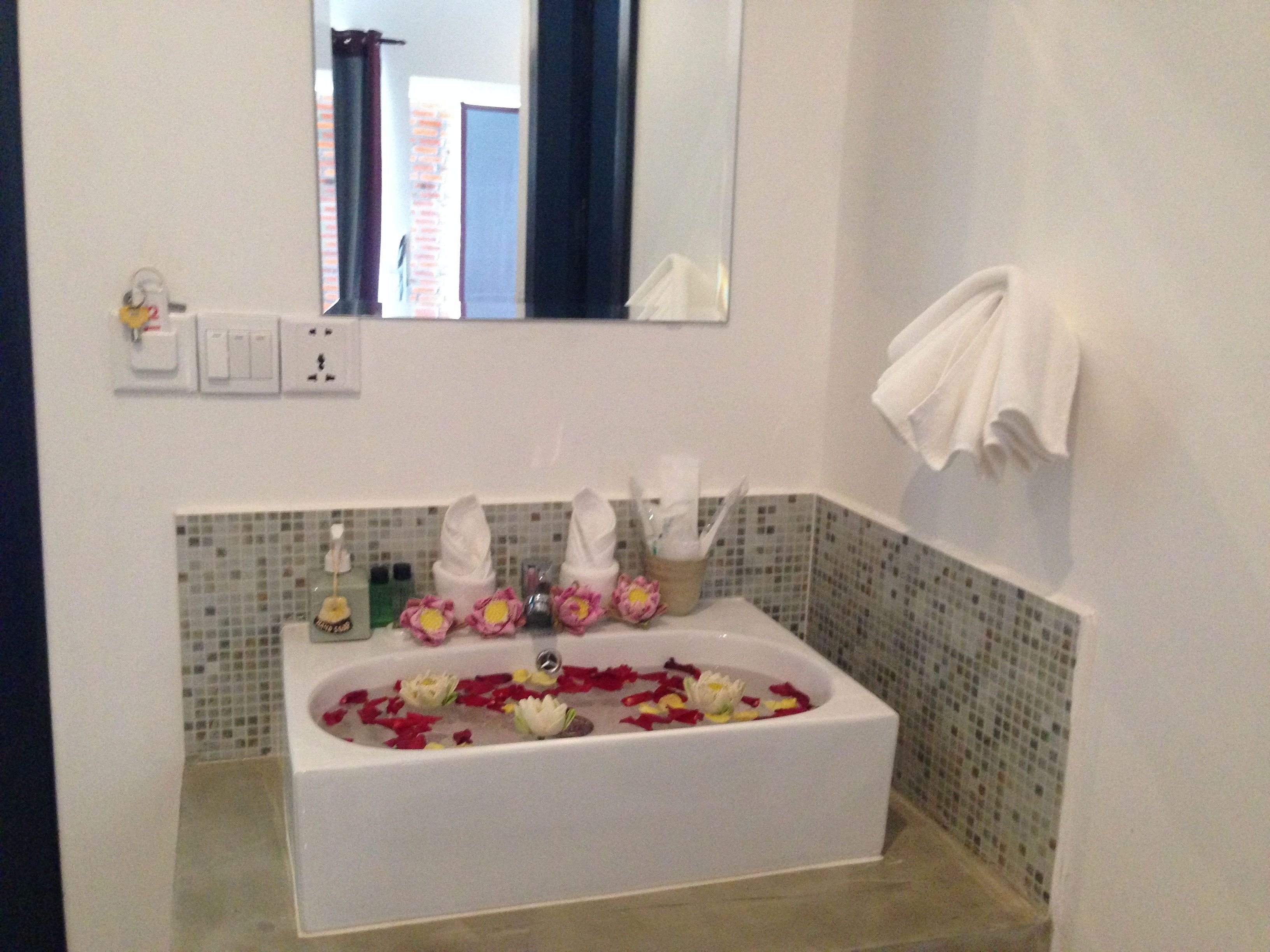 A bathroom with a white bathtub filled with rose petals and flowers, a mirror above the sink, and a towel rack on the wall.