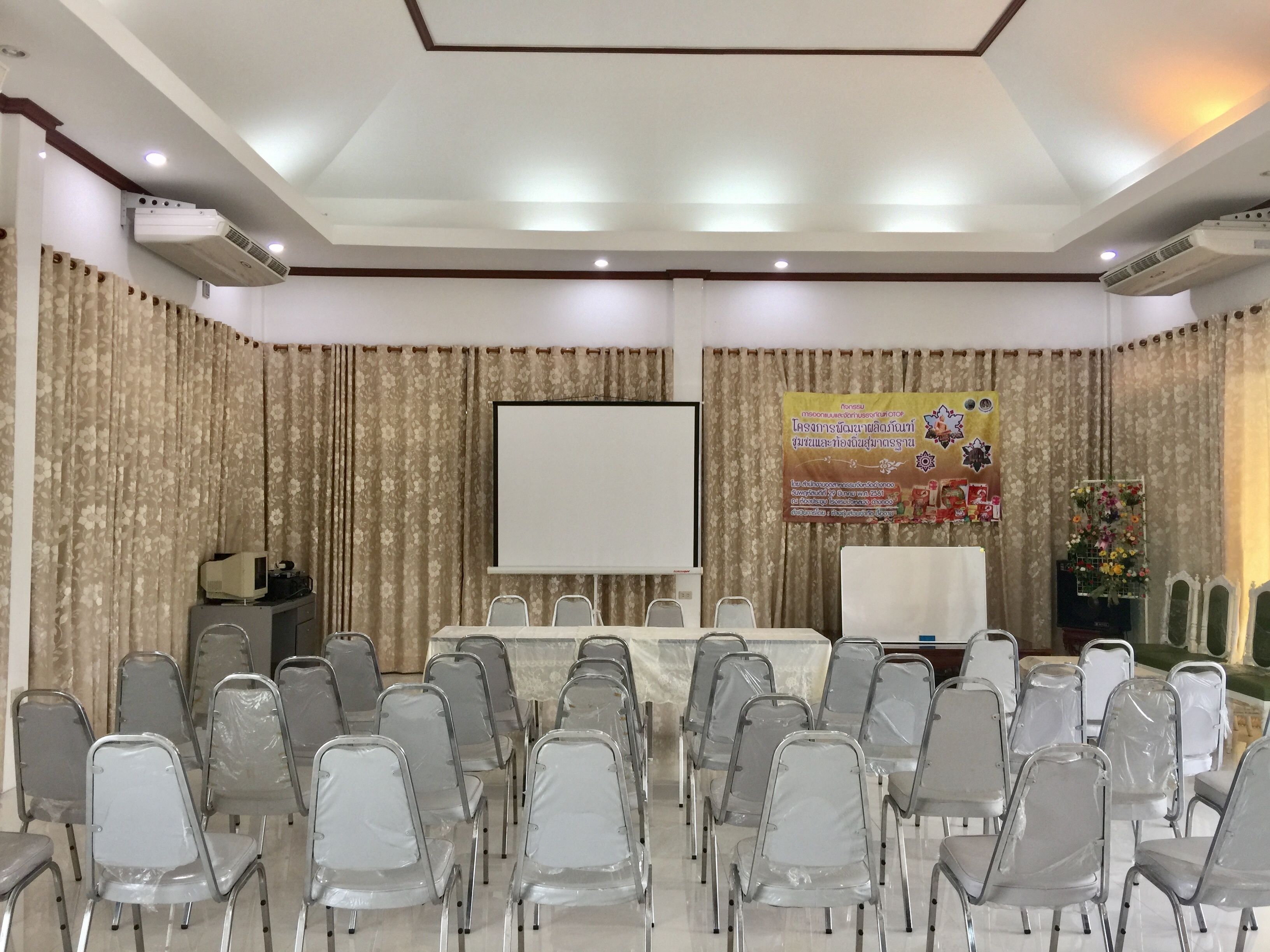 Uma sala de conferências com uma mesa branca, cadeiras de plástico transparente, uma tela de projetor e um banner na parede.