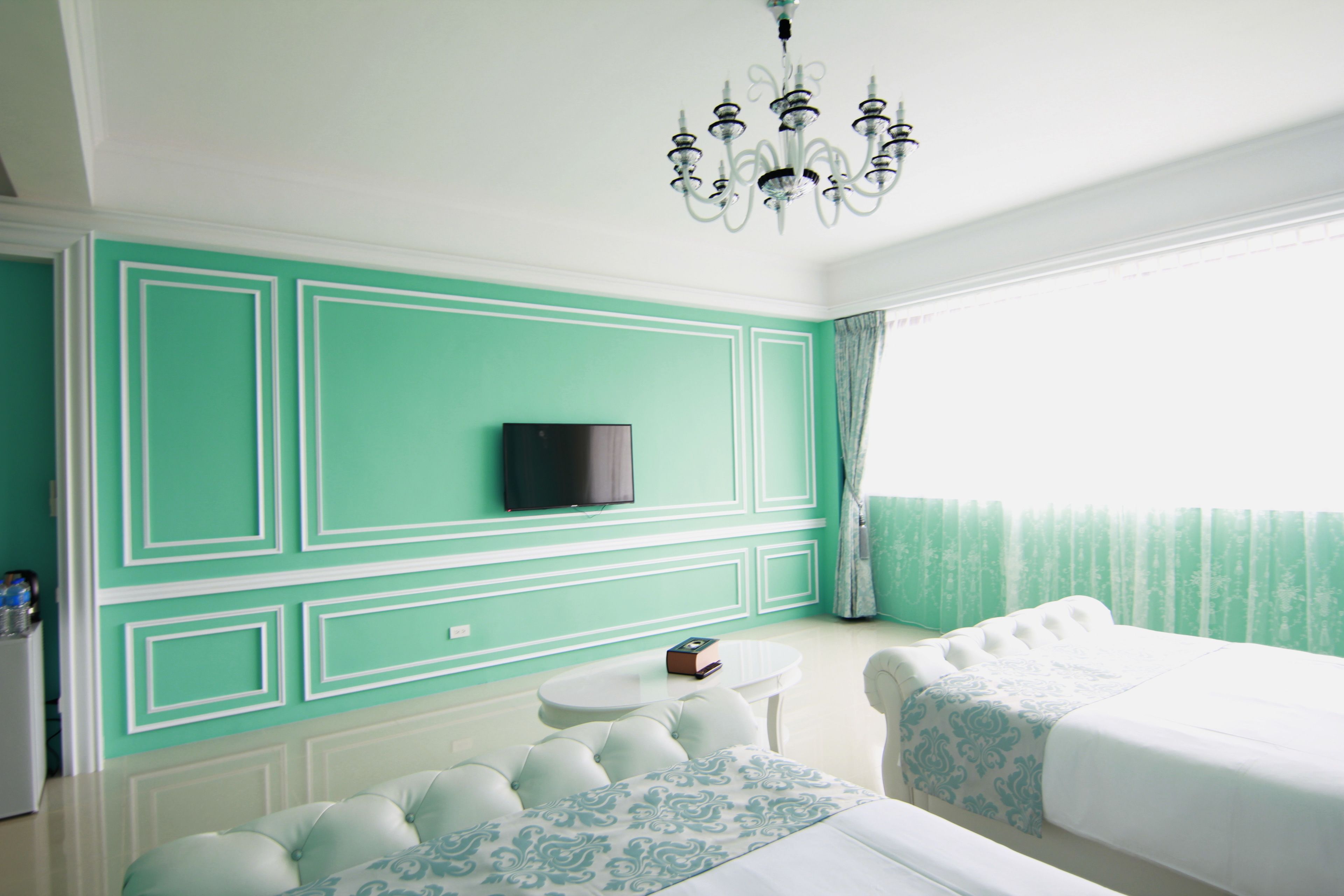 A room with teal walls, a flat-screen TV mounted above a white round table, a bed with a white headboard, and a chandelier.
