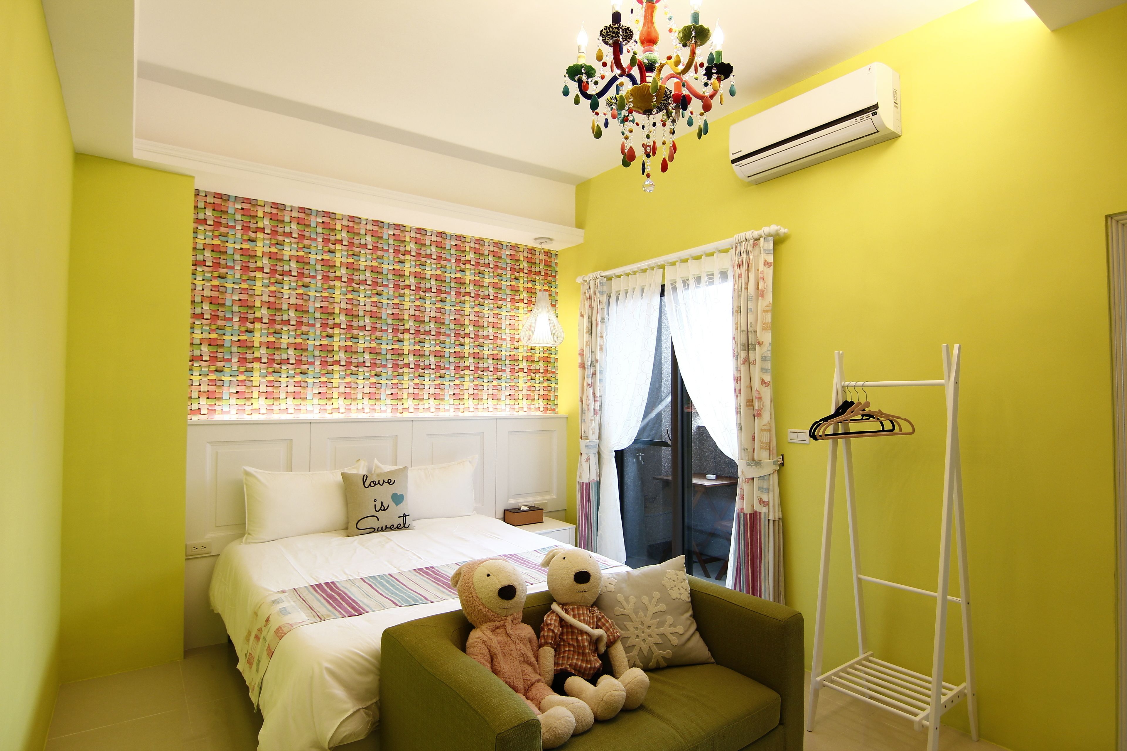 Quarto com cama, poltrona verde, dois ursinhos de pelúcia, ar-condicionado branco e janela com cortinas.