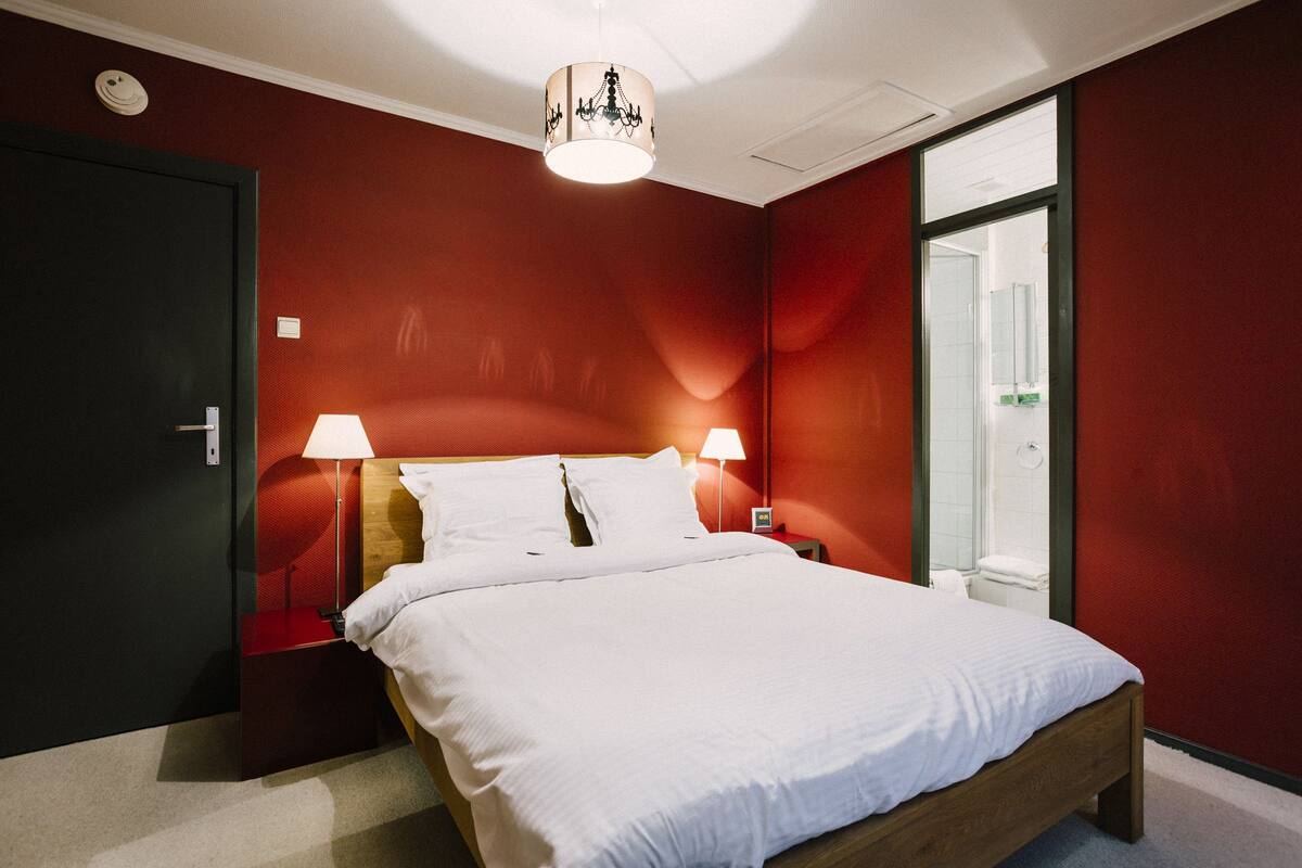 A modern hotel room with a large bed, two bedside lamps, a chandelier, and a glass door leading to a bathroom.