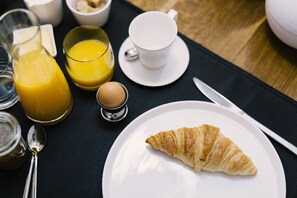 A breakfast setting with a croissant on a plate, a glass of orange juice, a cup of coffee, and a small bowl of sugar. - Tripel B (Bruges)