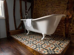 Une baignoire à pieds de couleur blanche, dans une pièce dont les murs sont en briques apparentes et dont le sol est recouvert de carreaux décoratifs.