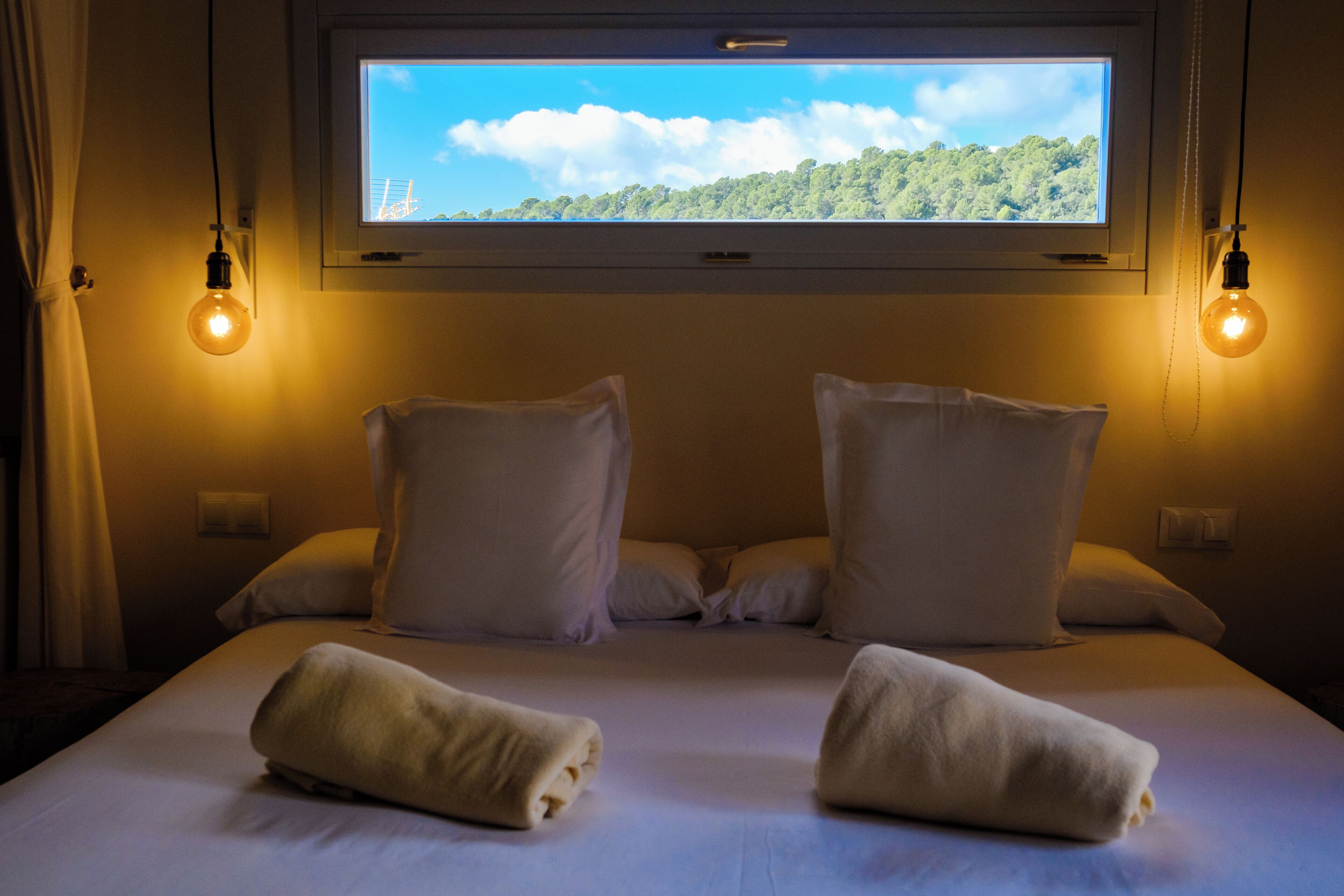 Habitación de hotel con una cama grande, dos almohadas, dos toallas y una ventana con vista a un bosque.