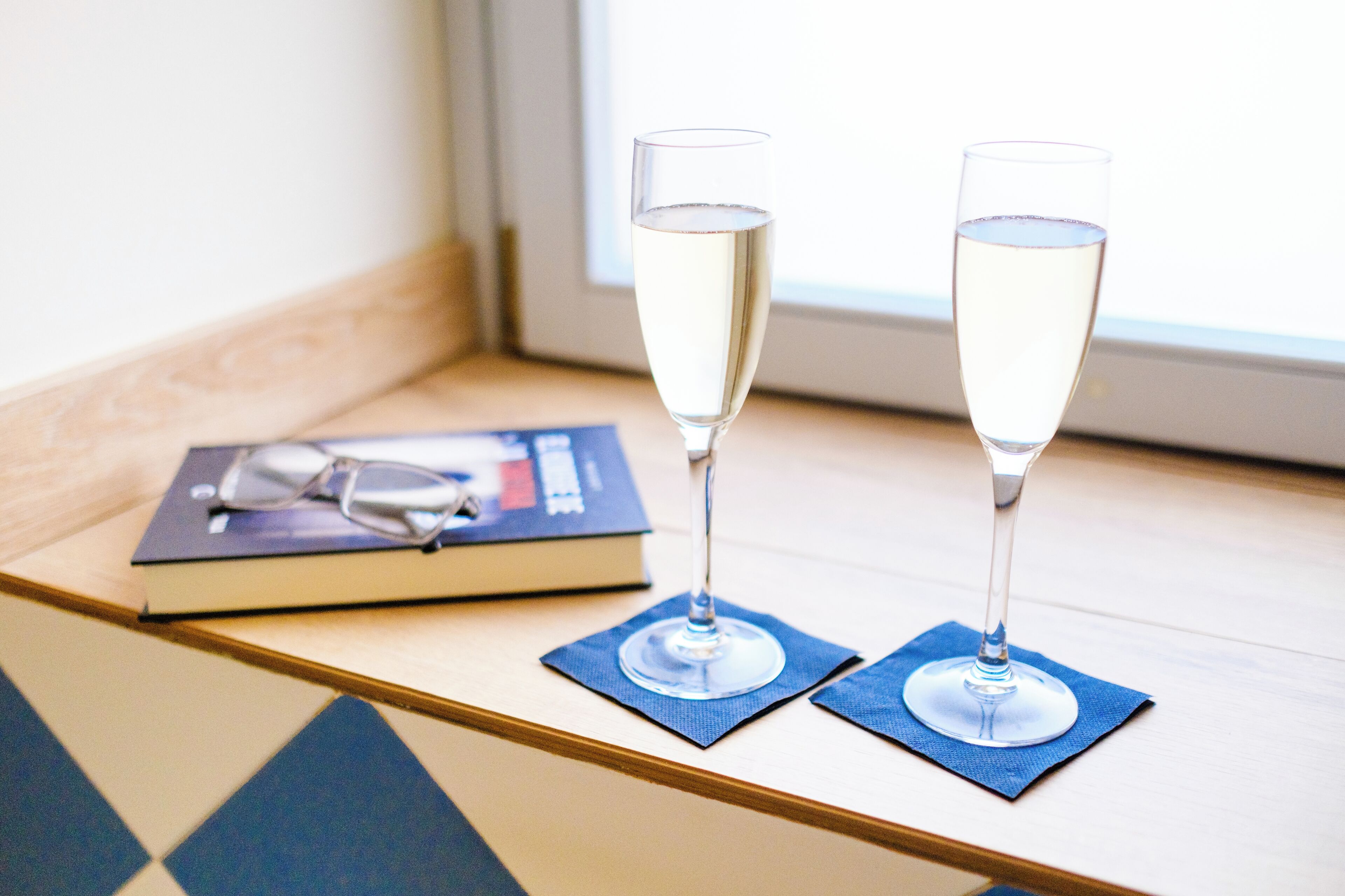 Zwei Sektgläser auf blauen Servietten auf einem Holztisch, neben ihnen ein Buch und Gläser.