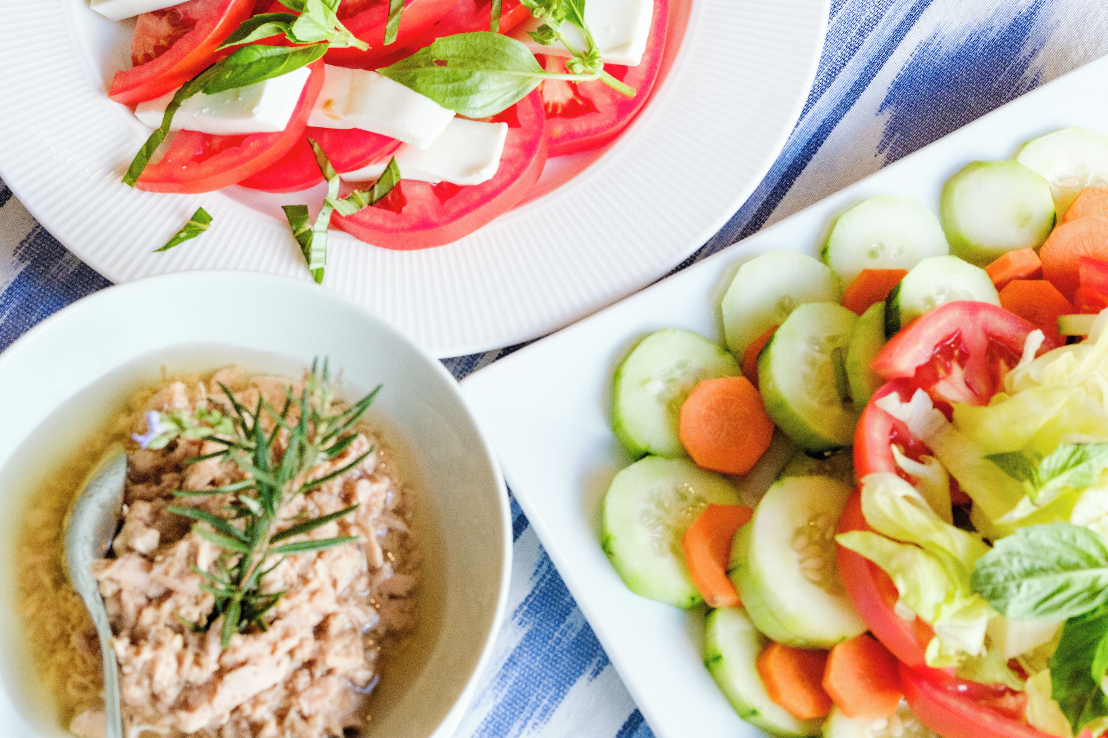 Ein Tisch mit drei Tellern Essen: ein Tomaten-Mozzarella-Salat, ein Gurken-Karotten-Salat und eine Schüssel Thunfischsalat mit Kräutern.