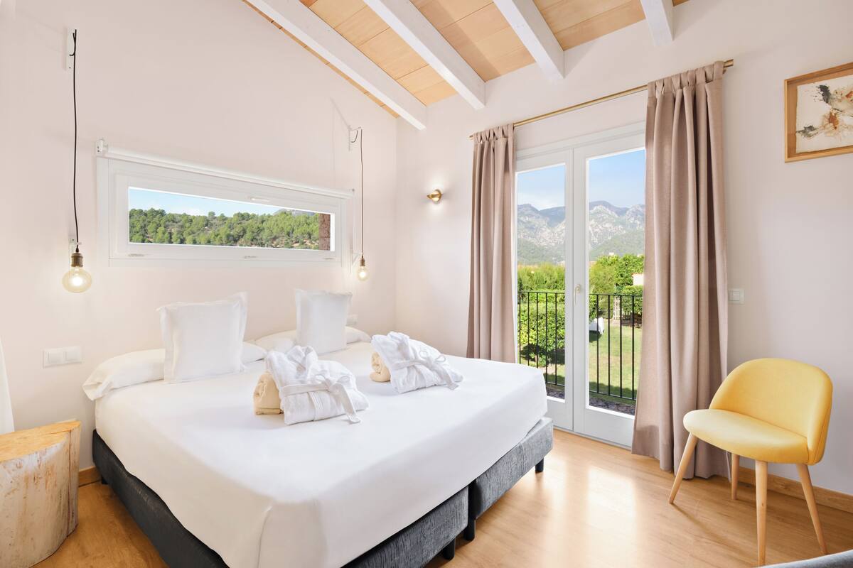 A bedroom with a large bed, a window with a view of greenery, a balcony with a chair, and a wooden ceiling.