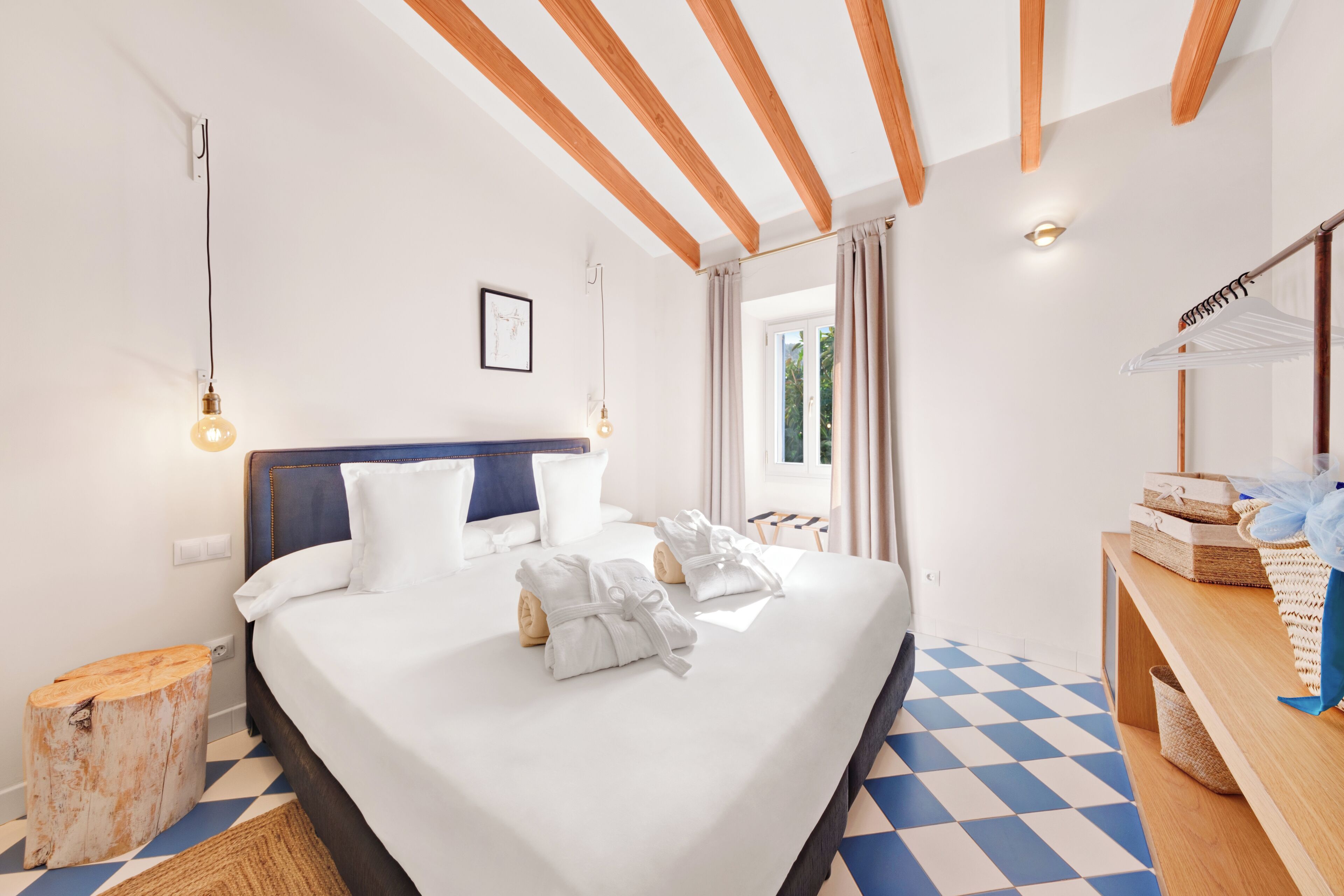 A hotel room with a large bed, wooden beams on the ceiling, a window with curtains, and a wooden side table with baskets.