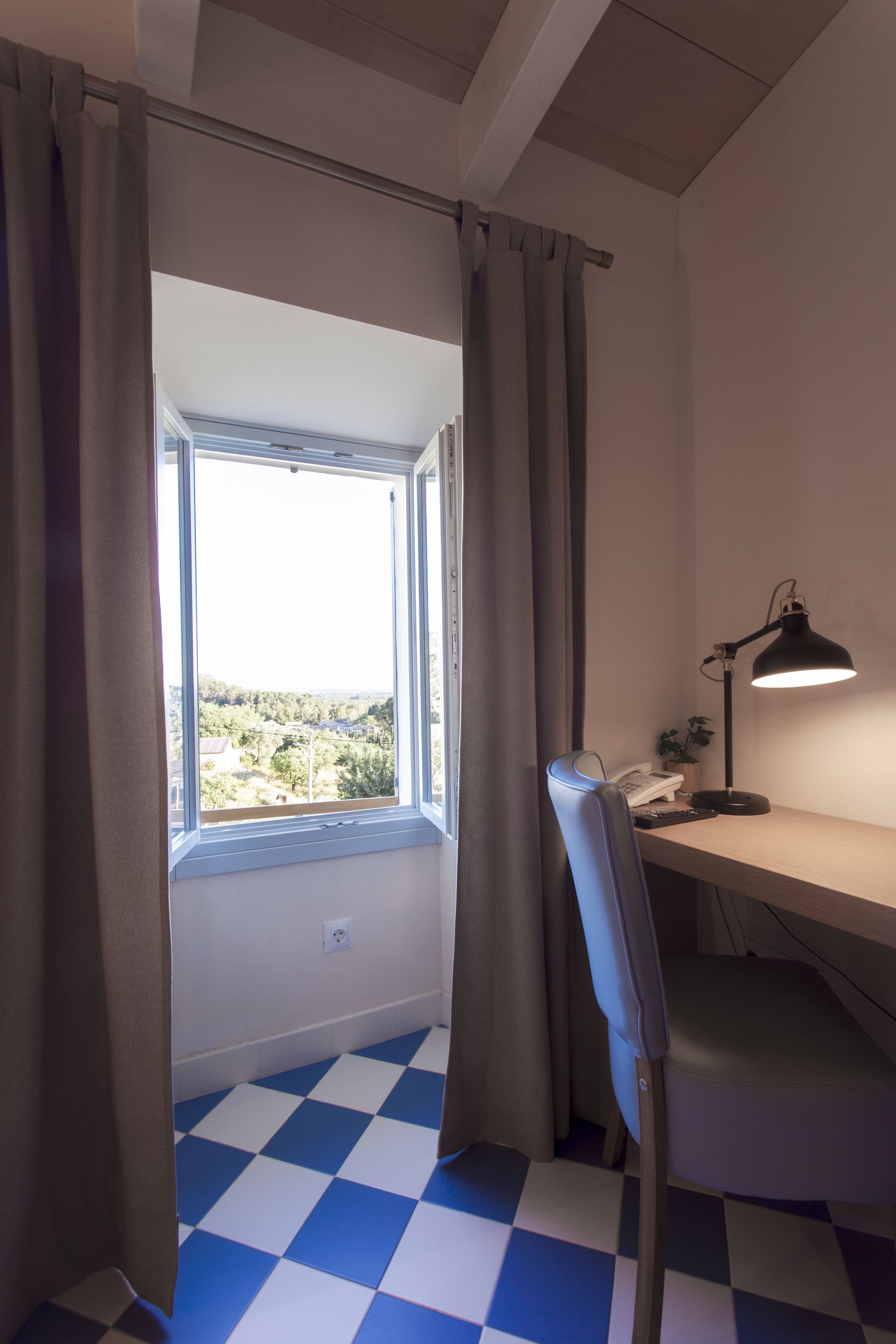 A room with a checkered floor, a desk with a lamp, a chair, and a window with curtains.