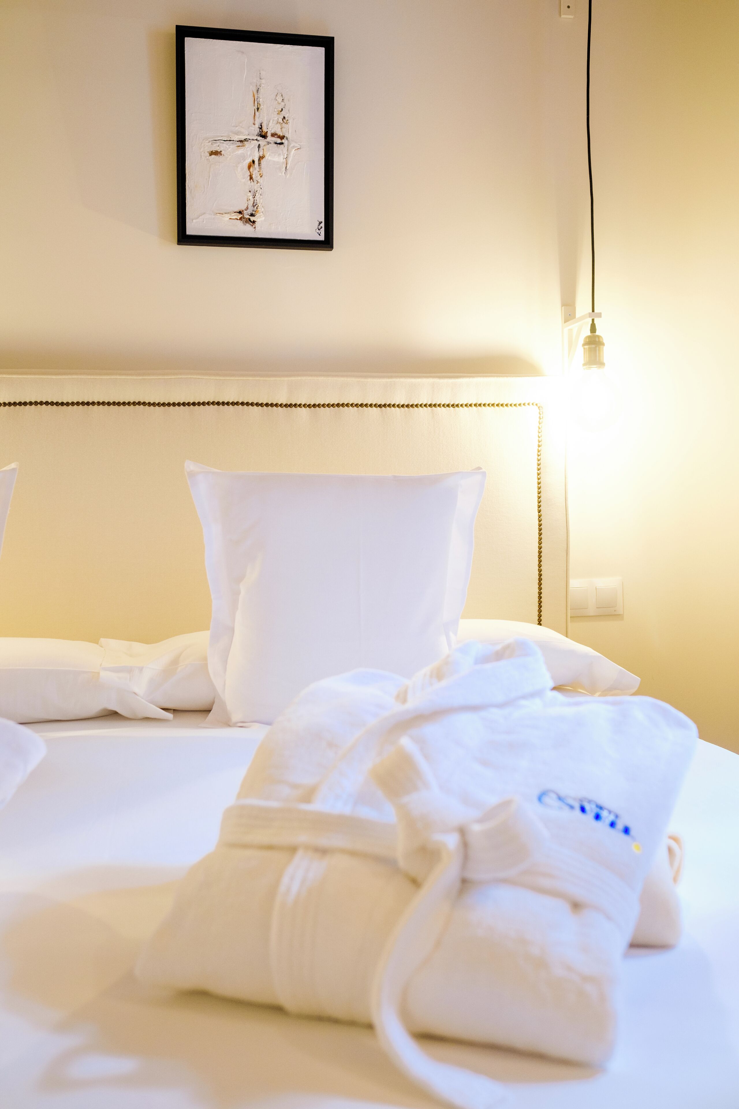 Habitación de hotel con una cama bien hecha, ropa blanca, una obra de arte enmarcada en la pared y una lámpara de noche.