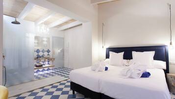 A hotel room with a checkered floor, a large bed with white bedding and multiple pillows, a dark headboard, and a bathroom with a glass shower enclosure and a chandelier.