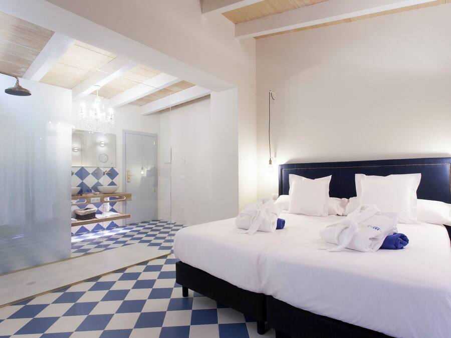 A hotel room with a checkered floor, a large bed with white bedding and multiple pillows, a dark headboard, and a bathroom with a glass shower enclosure and a chandelier.