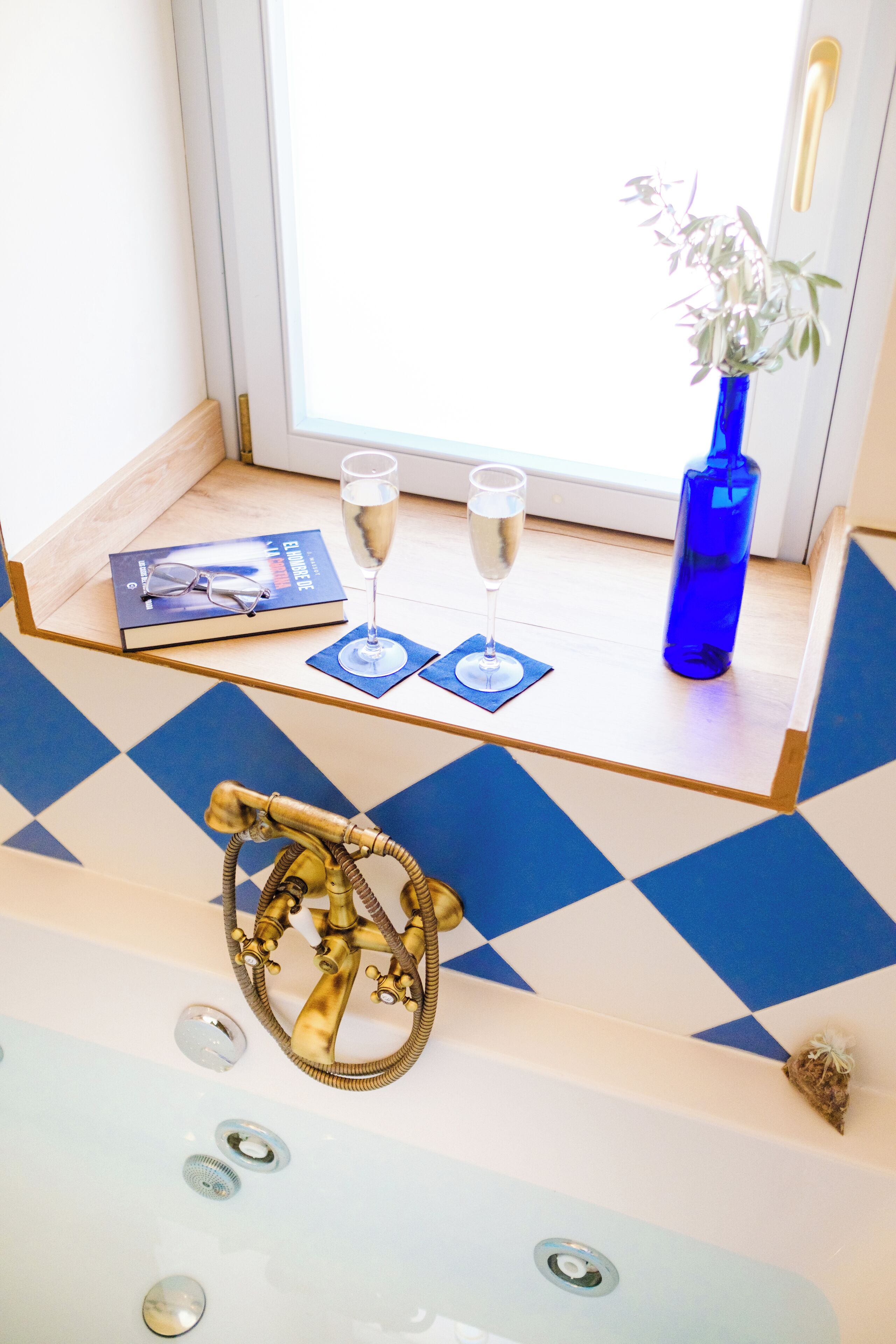 Ett rum med ett schackrutigt golv, ett fönster med utsikt, en blå vas med en växt, en bok och två glas champagne på ett litet bord.