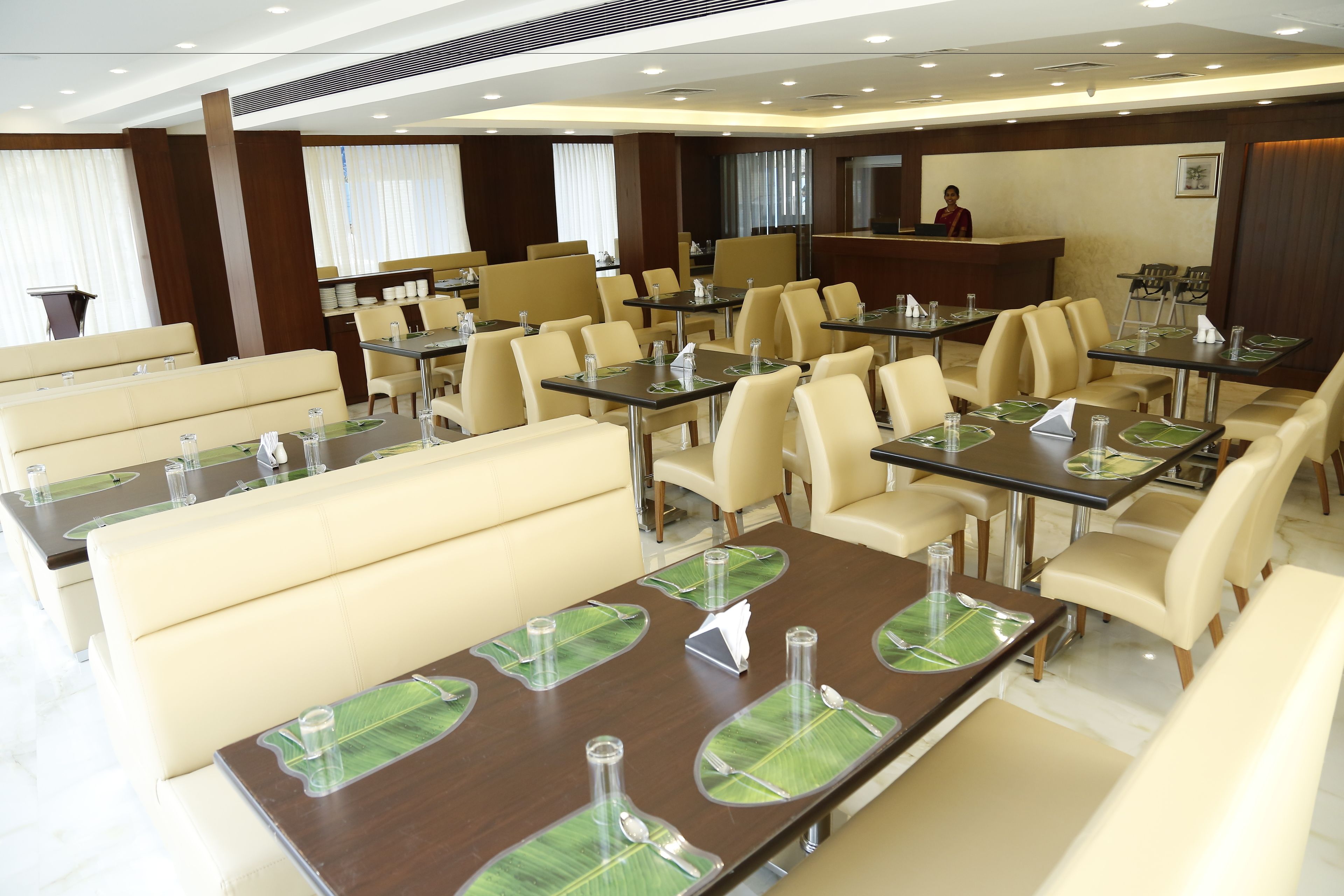 Un comedor de hotel con varias mesas y cabinas, una recepción y un amplio ventanal con cortinas.