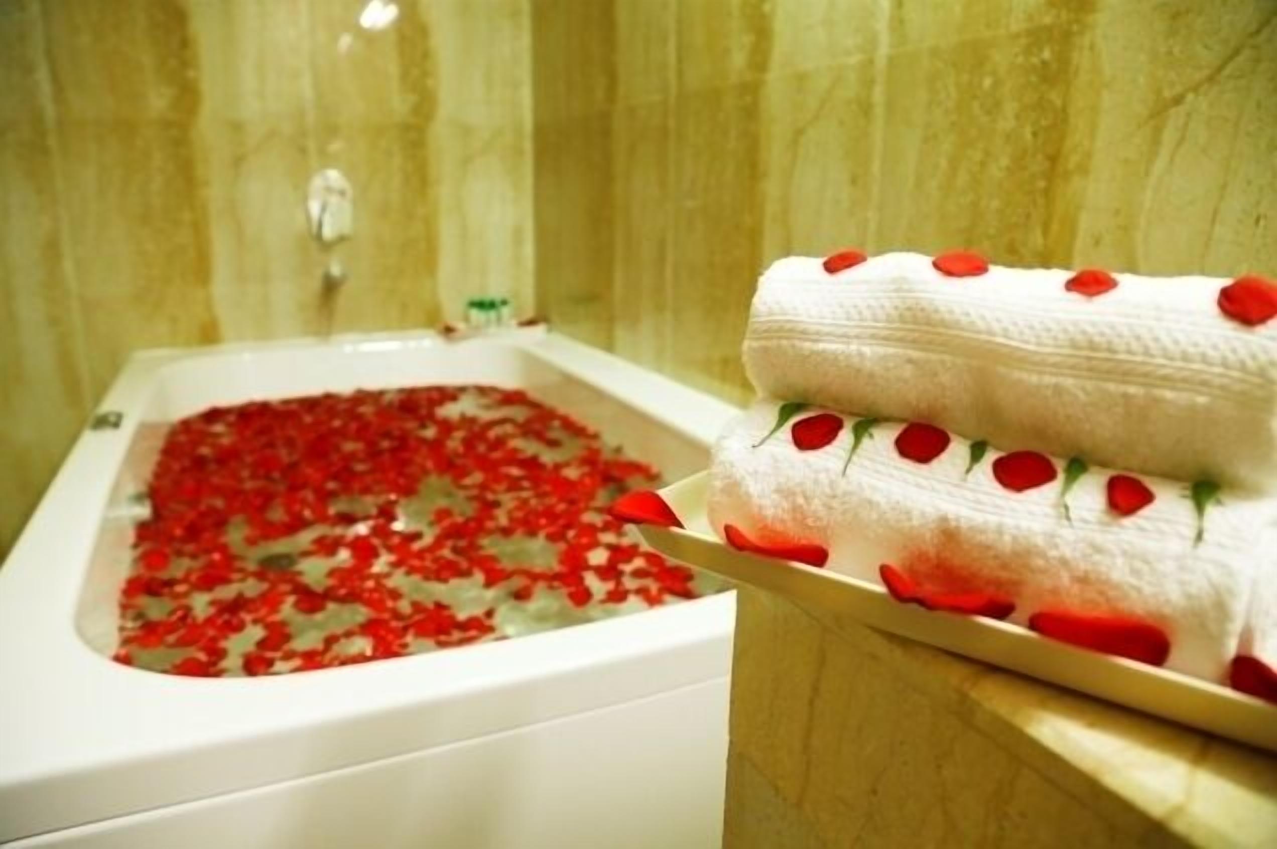 A bathtub filled with water and rose petals, with a towel with rose petals on a nearby surface.