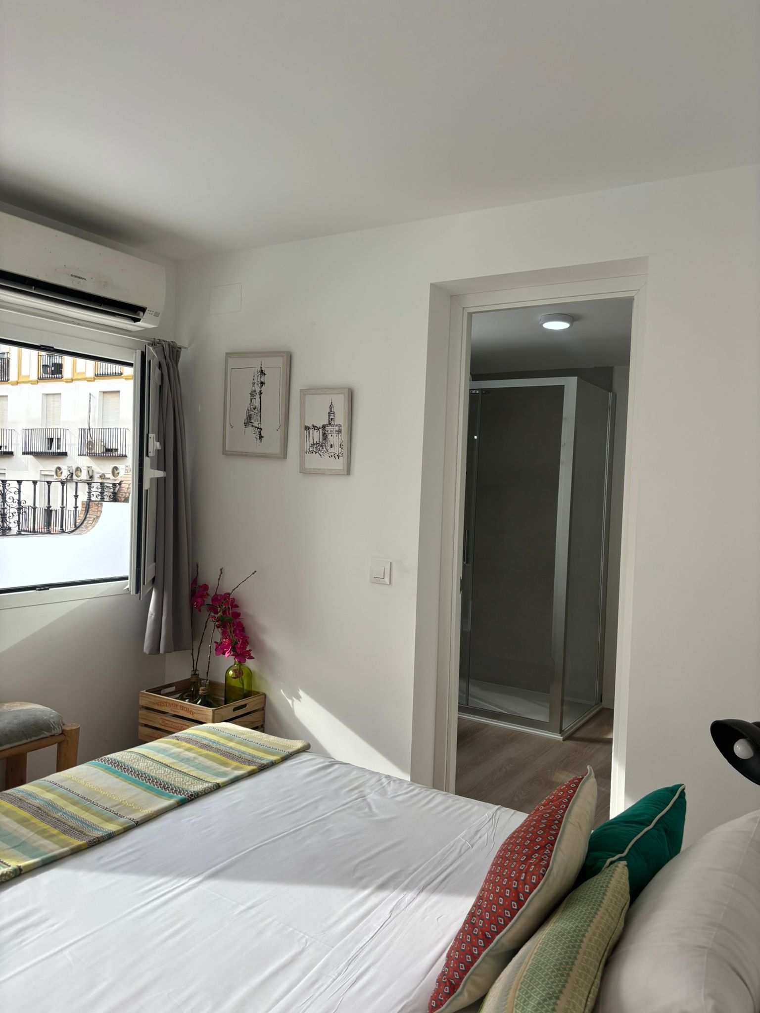 A bedroom with a bed, a window with curtains, a door leading to another room, and two framed pictures on the wall.