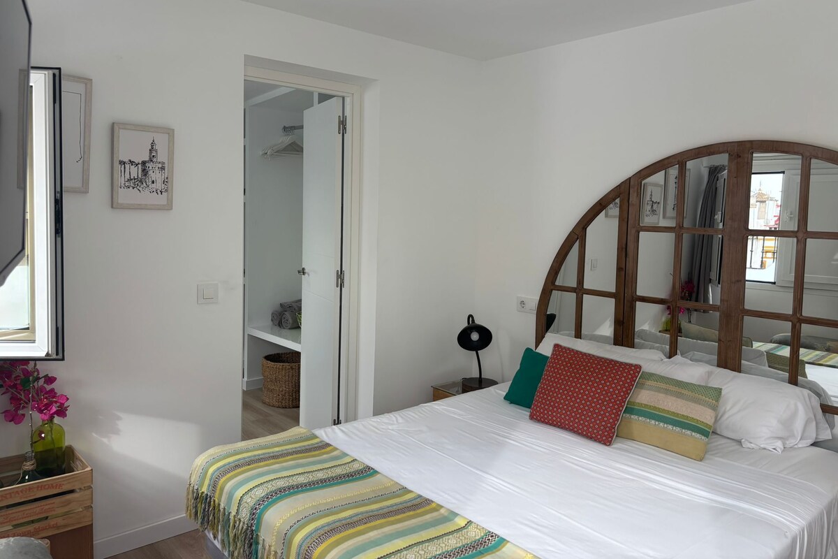 A bedroom with a bed, a wooden-framed mirror, a bedside lamp, and a view of a hallway.