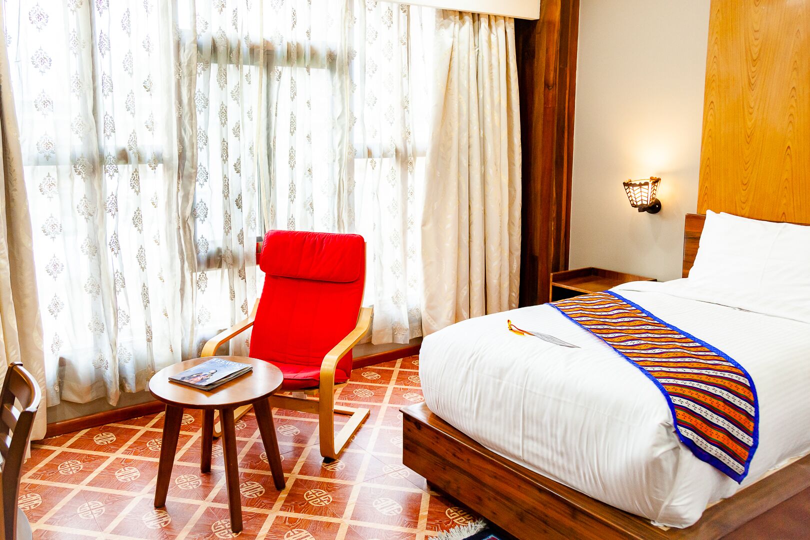 Una habitación de hotel con una cama, un sillón rojo, una mesita y cortinas.