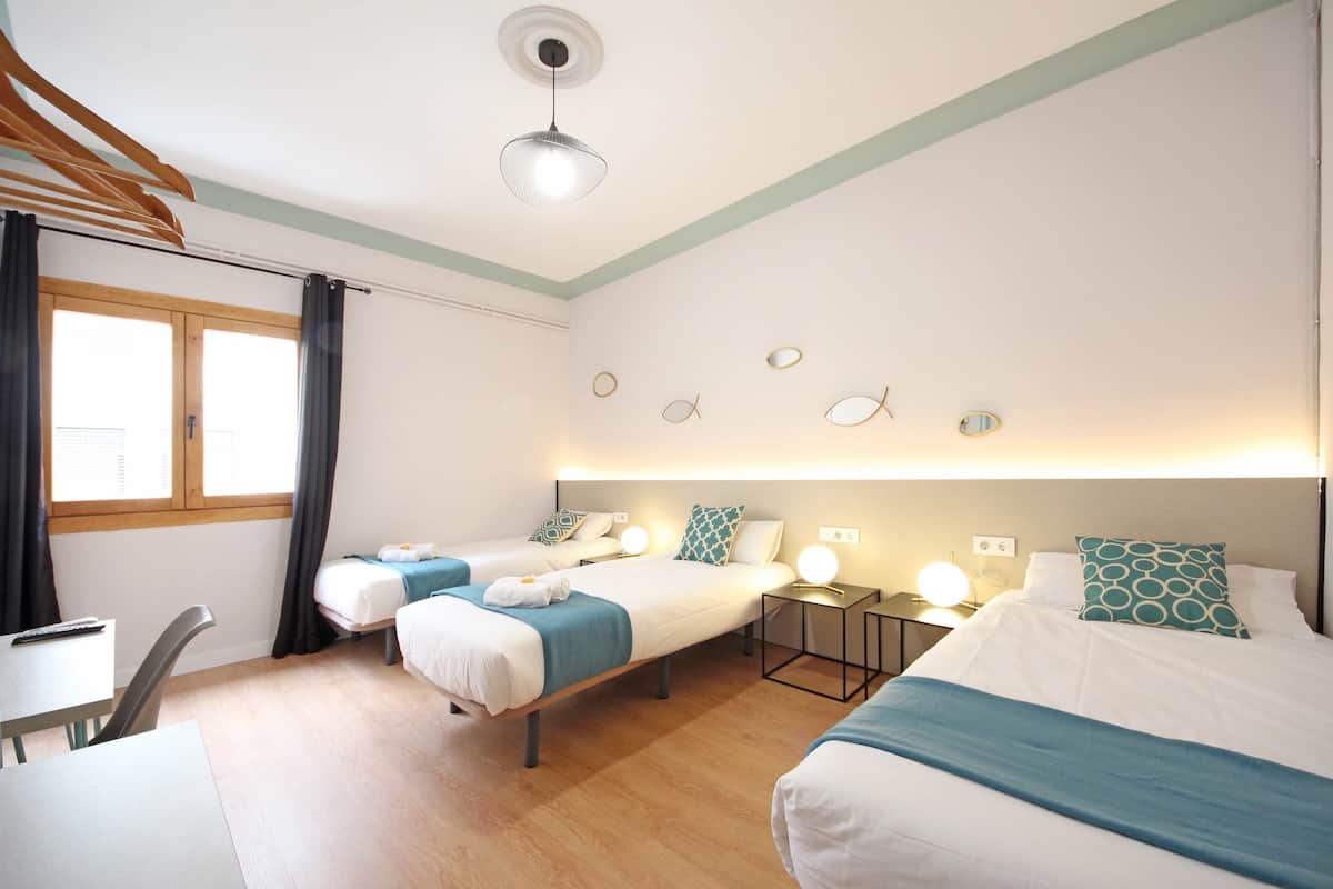 Une chambre d’hôtel avec deux lits, un bureau et une chaise. Il y a des rideaux à la fenêtre, un luminaire au plafond et des lampes fixées au mur.