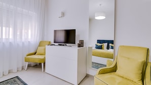 Ein modernes Zimmer mit Flachbildfernseher, einer weißen Kommode, zwei gelben Sesseln, einem großen Spiegel und einem Fenster mit transparenten Vorhängen.