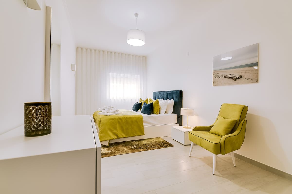 A modern bedroom with a bed, a green armchair, a bedside table, and a painting on the wall.