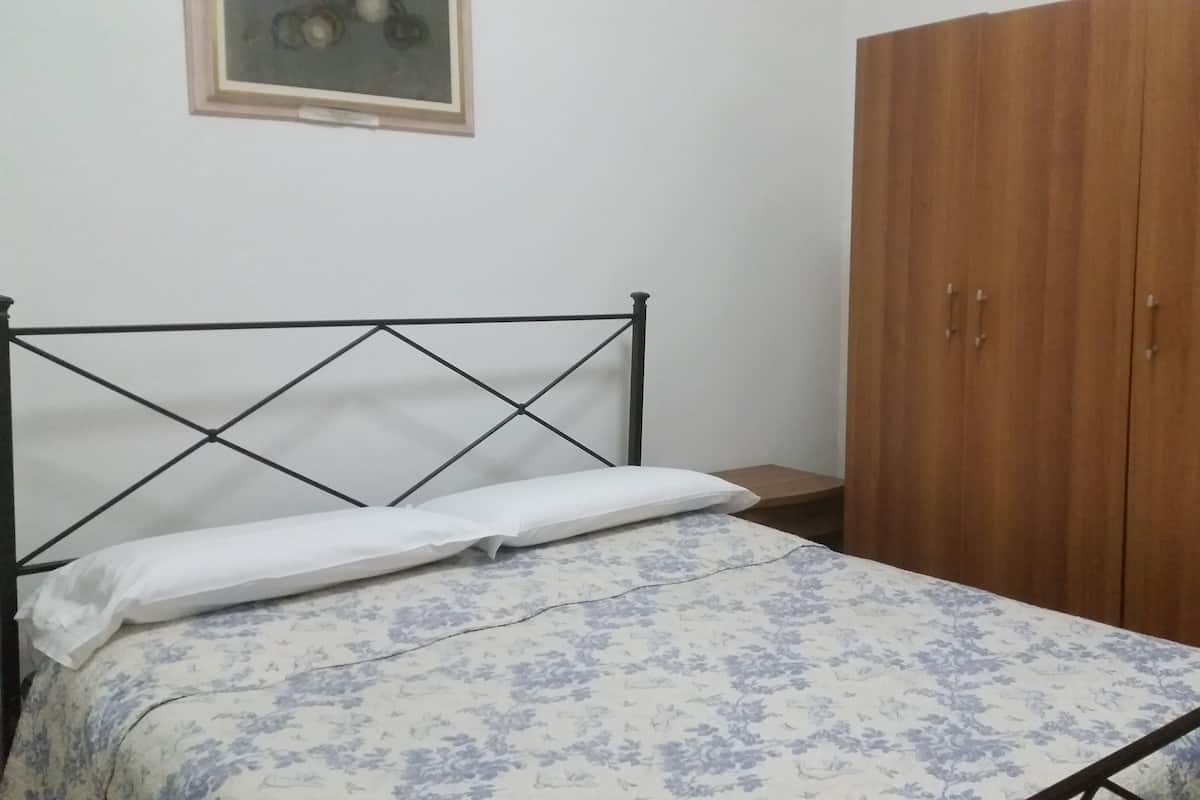 Een bed met een bloemenprint deken en een wit kussen, een houten garderobe en een klein nachtkastje.