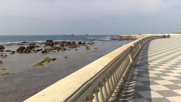 A coastal promenade with a checkered walkway, a rocky beach, and a distant ship.