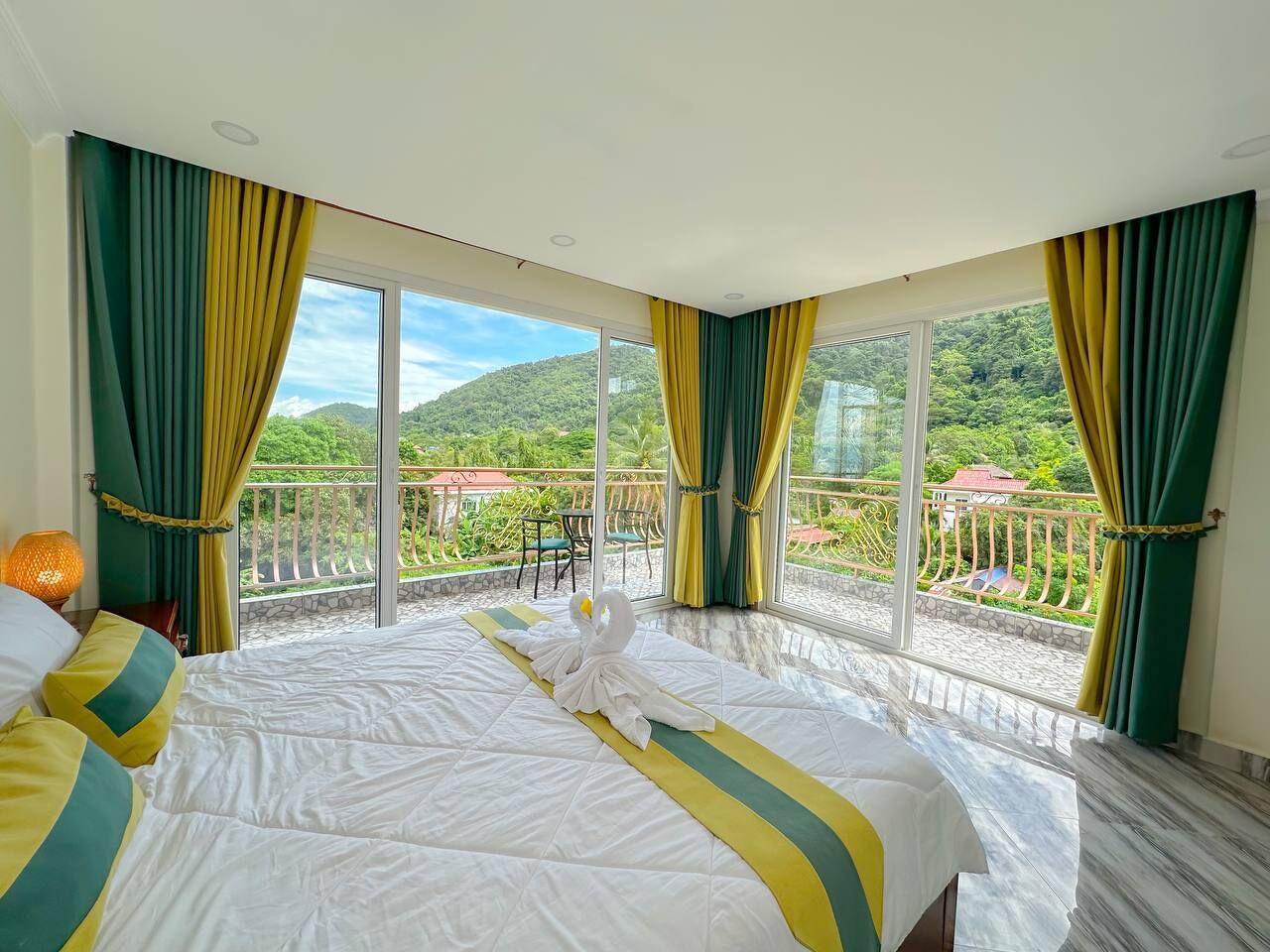 A hotel room with a large bed, two windows with curtains, a balcony with chairs, and a view of greenery.