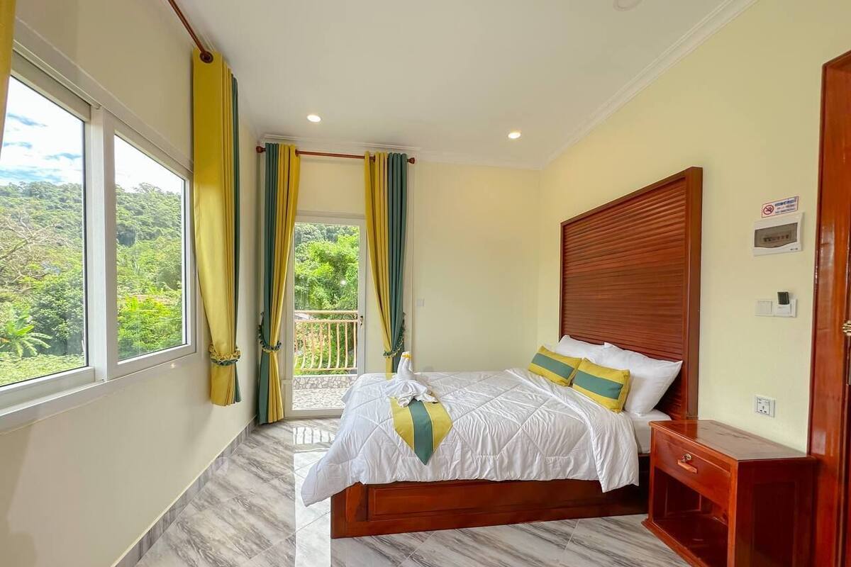Una camera da letto con un letto, una testiera in legno, un comodino, una finestra con tende e vista su piante.