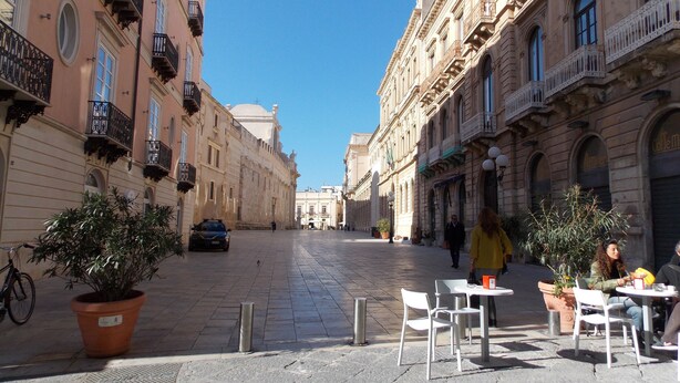Una calle adoquinada flanqueada por edificios históricos, mesas al aire libre y macetas con plantas.