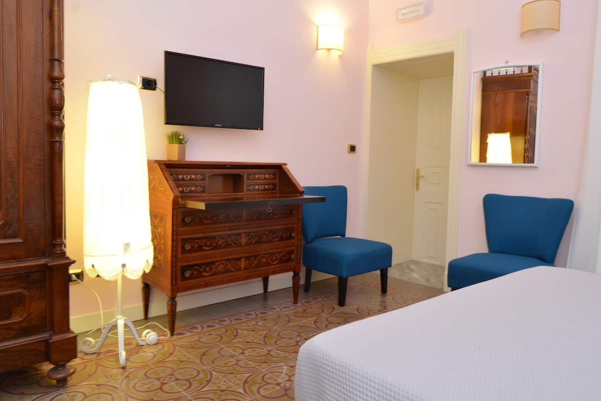 A hotel room with a bed, a wooden dresser, a television, two blue chairs, and a patterned floor.