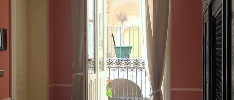 Una habitación con un gran ventanal abierto que da a un balcón con vistas, dos sillones tapizados, suelo de baldosas y un aire acondicionado instalado en la pared.