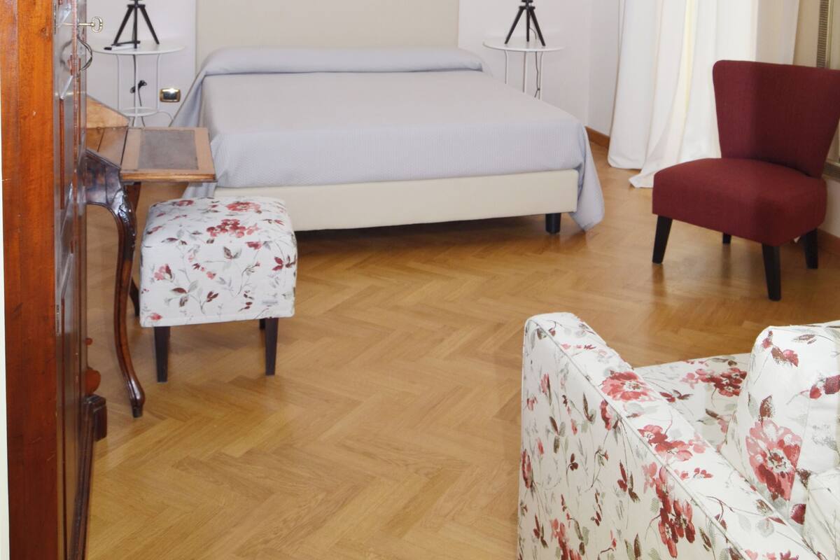 A bedroom with a bed, two bedside lamps, a wooden chair, a small table, and a red armchair.