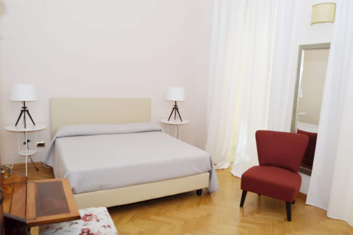 A hotel room with a bed, two bedside lamps, a red chair, a small table, and a mirror.