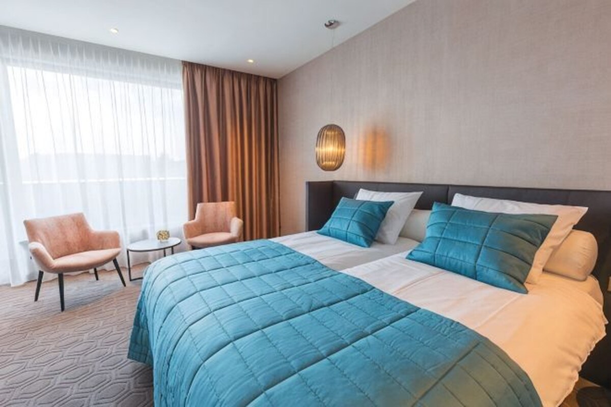 A hotel room with a large bed, two armchairs, a small table, and a view of the outdoors through sheer curtains.