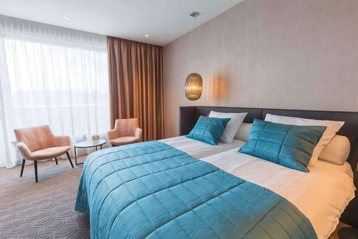 A hotel room with a large bed, two armchairs, a small table, and a view of the outdoors through sheer curtains.