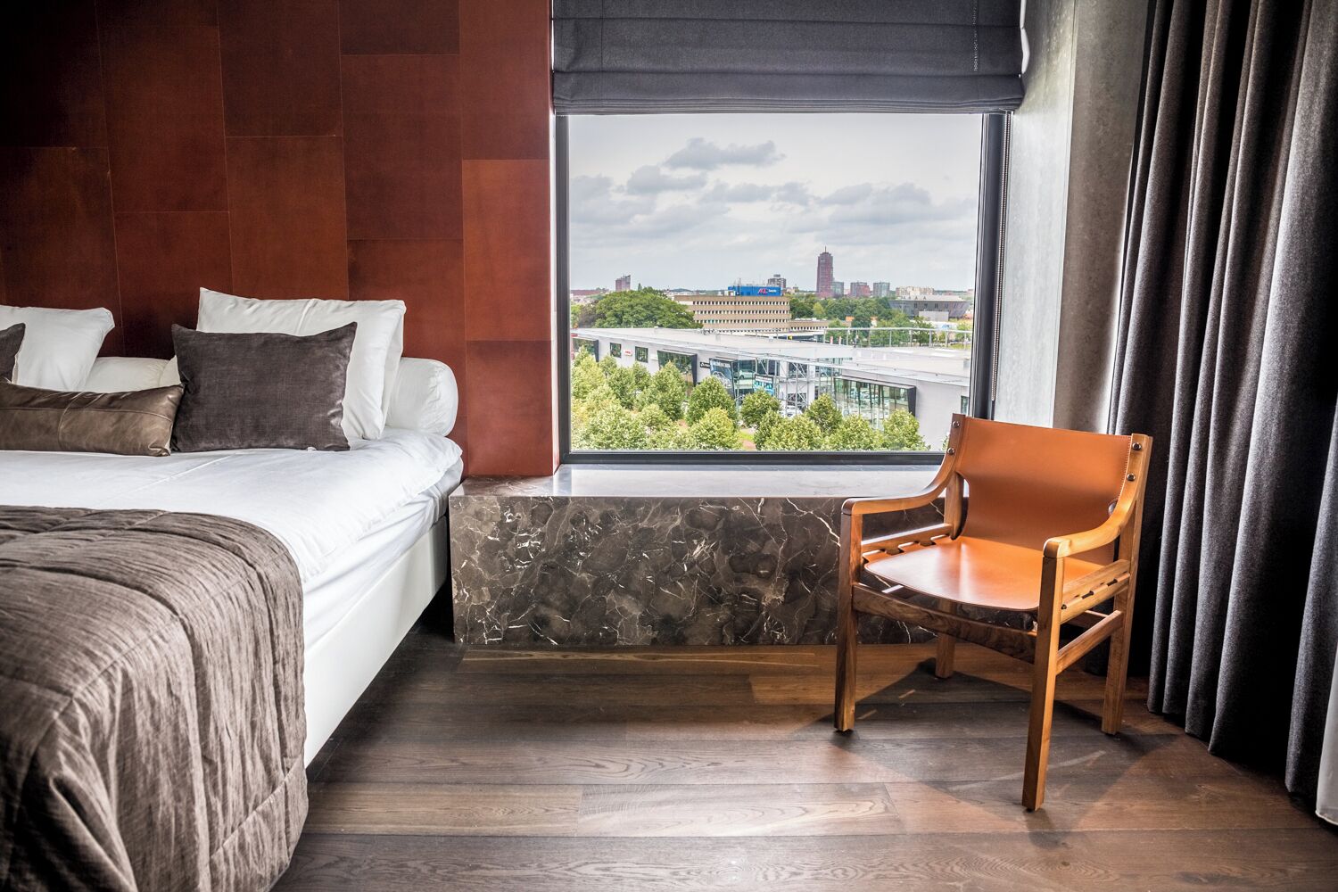 A hotel room with a bed, a chair, a window with a city view, and a marble accent wall.
