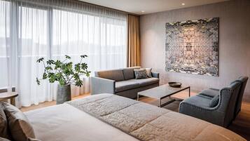 A modern hotel room with a large bed, a sofa, a chair, a coffee table, and a large decorative wall piece.