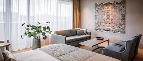 Een moderne hotelkamer met een groot bed, een slaapbank, een fauteuil, een bijzettafeltje en een groot decoratief wandobject.
