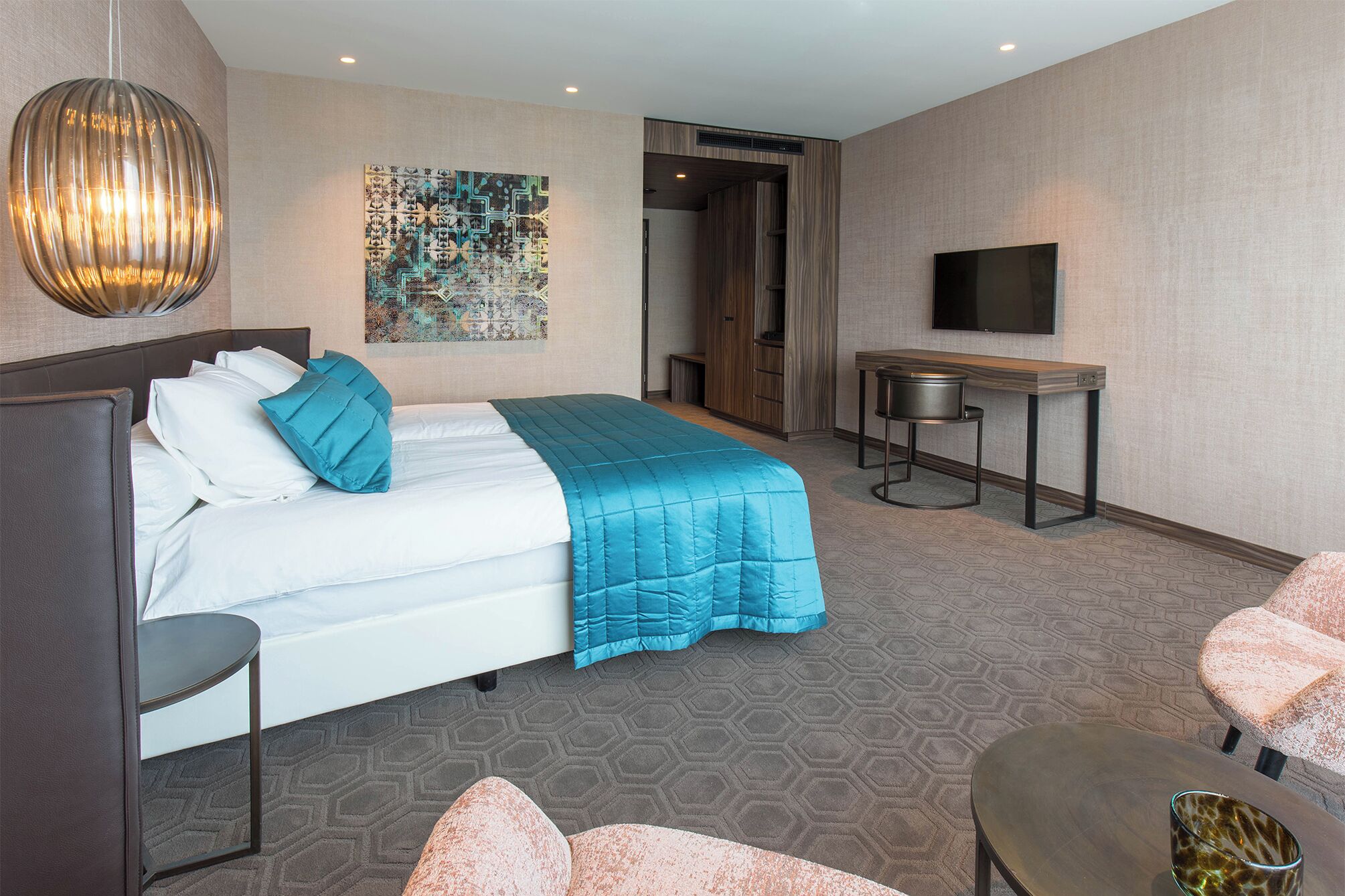 A hotel room with a bed, a desk, a chair, a TV, and a wall art piece.