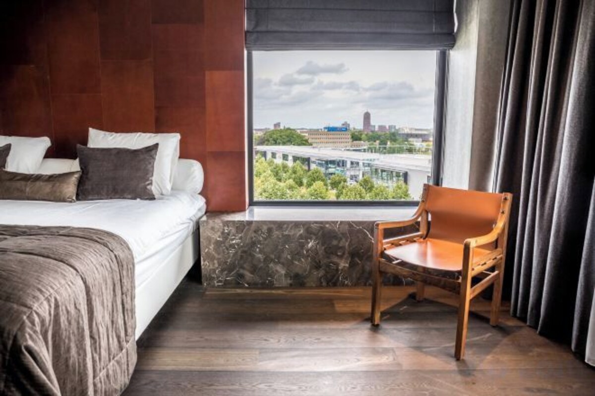 A hotel room with a bed, a chair, a window with a city view, and a curtain.
