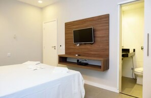 Un dormitorio con cama, un televisor montado en un panel de madera y un baño visible a través de una puerta abierta.