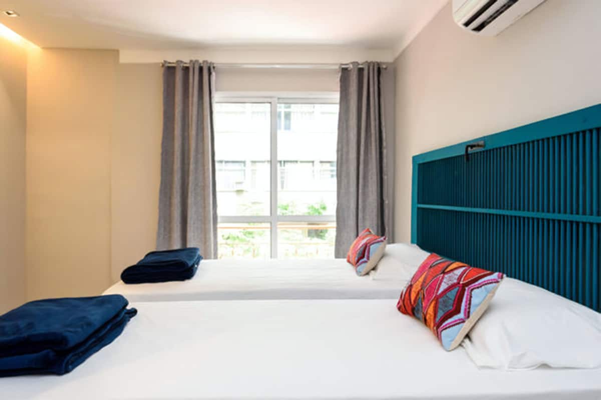 Quarto de hotel com duas camas, janela com cortinas, cabeceira verde-água e vista para o verde lá fora.